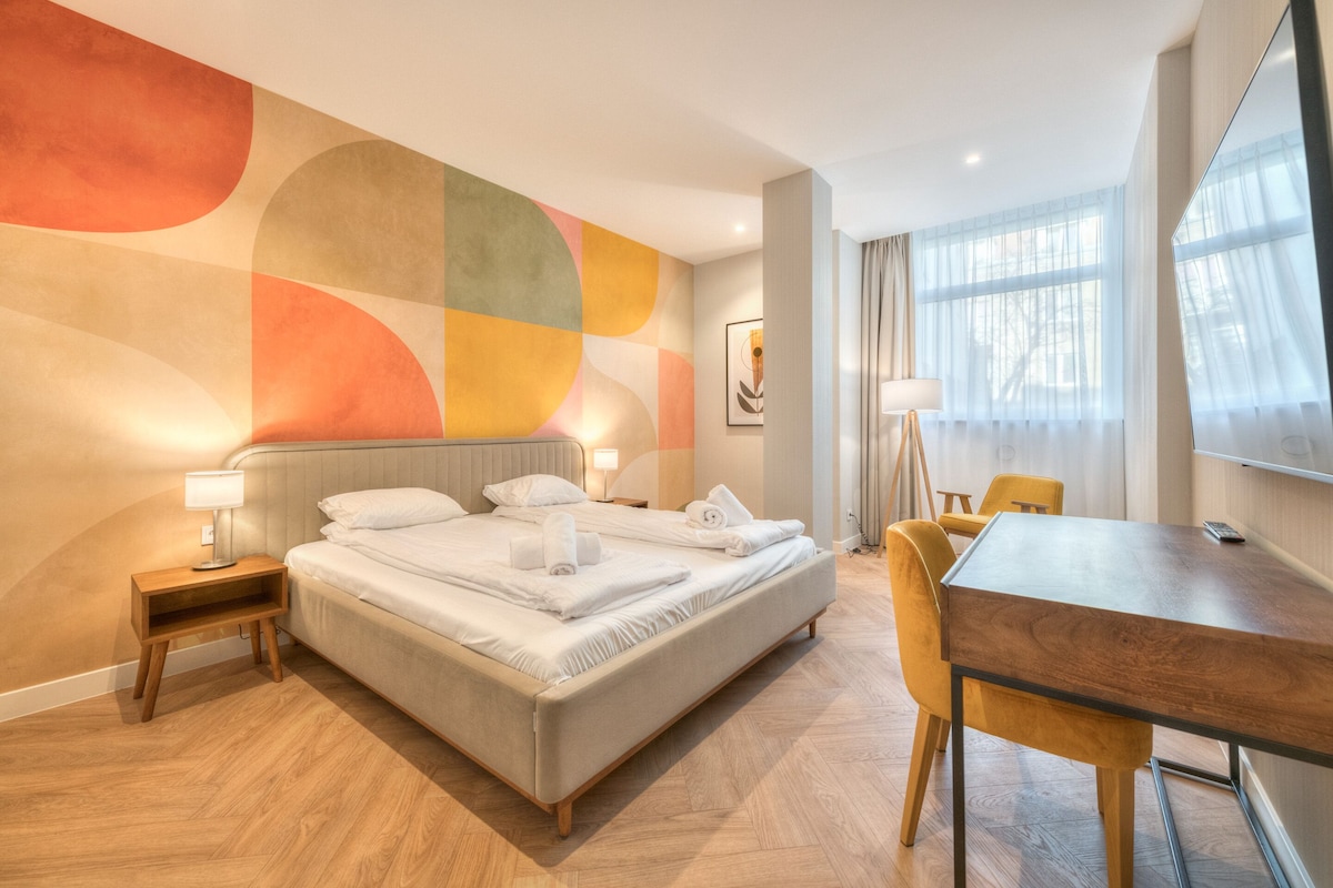 Una habitación de hotel con una cama, mesitas de noche, un escritorio y un ventanal amplio.