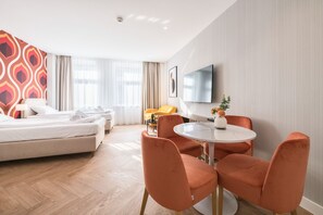 Ett hotellrum med en säng, två orangefärgade stolar, ett litet runt bord, en tv och ett fönster med gardiner.