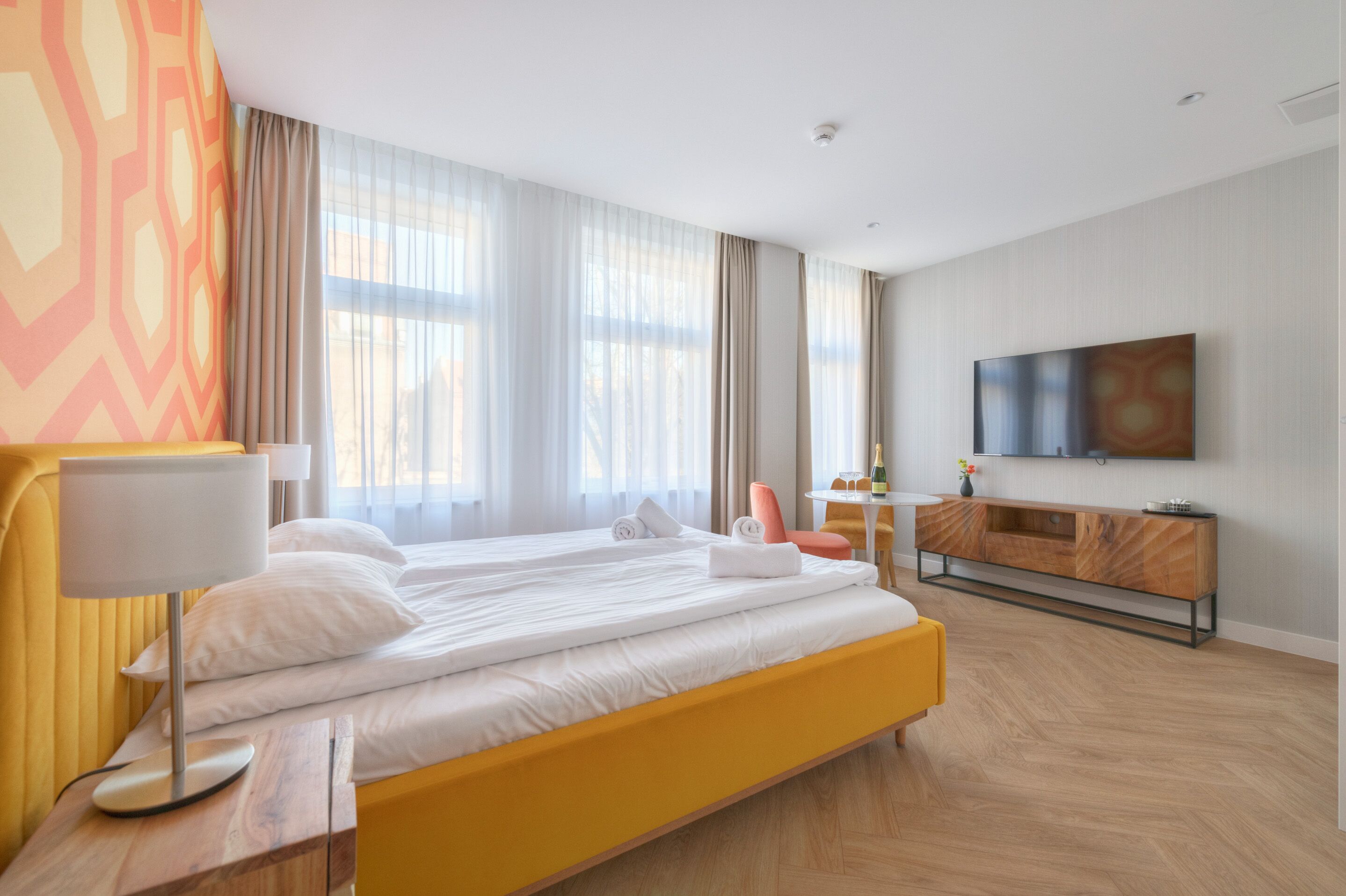 A hotel room with a large bed, a bedside table with a lamp, a small table with a chair, a TV mounted on the wall, and a large window with curtains.