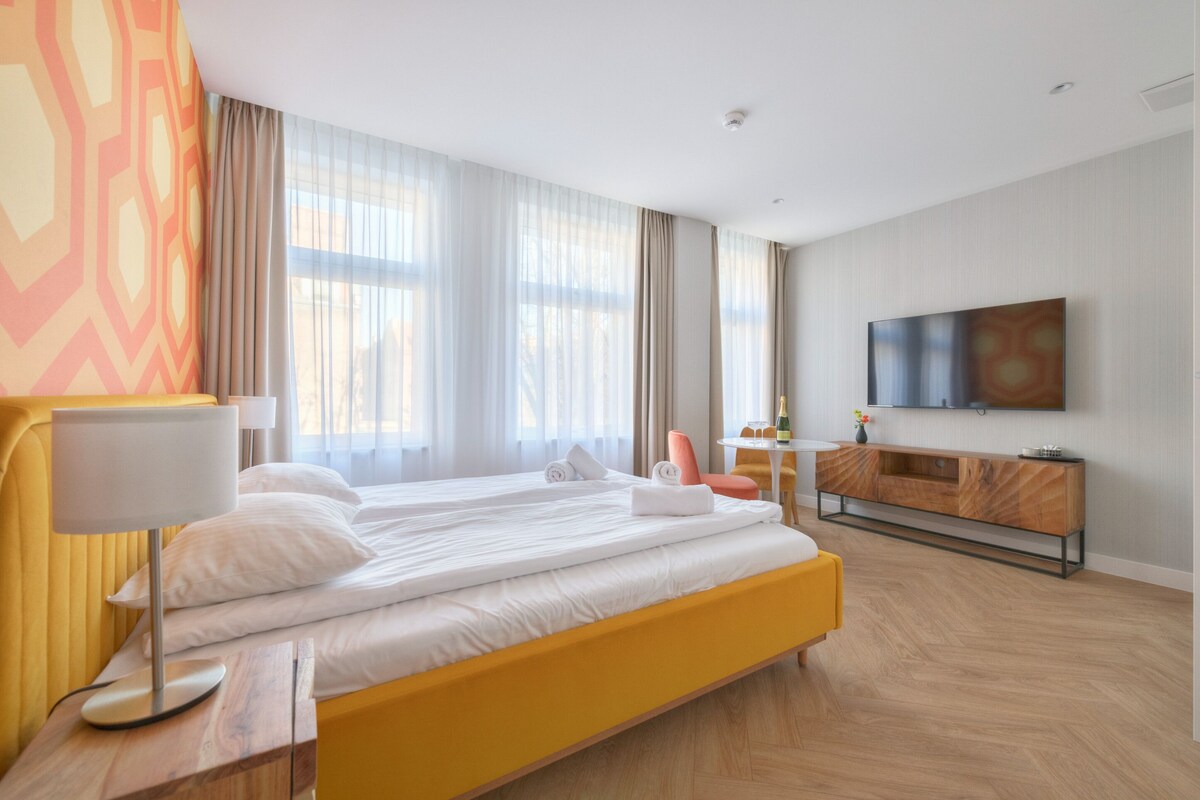 Habitación de hotel con una cama grande, una mesita de noche con lámpara, una pequeña mesa con una silla, un televisor montado en la pared y un amplio ventanal con cortinas.