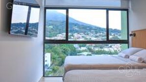 A bedroom with a large window offering a view of a cityscape and mountains. - Cozy, Modern APT @ Col. Escalon - Pool, Gym, A/C (San Salvador)