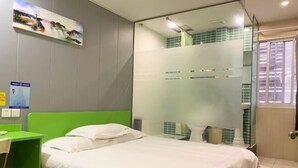 Quarto de hotel com cabeceira verde, cama com roupa de cama branca, divisória de vidro separando o espaço e janela com persianas.