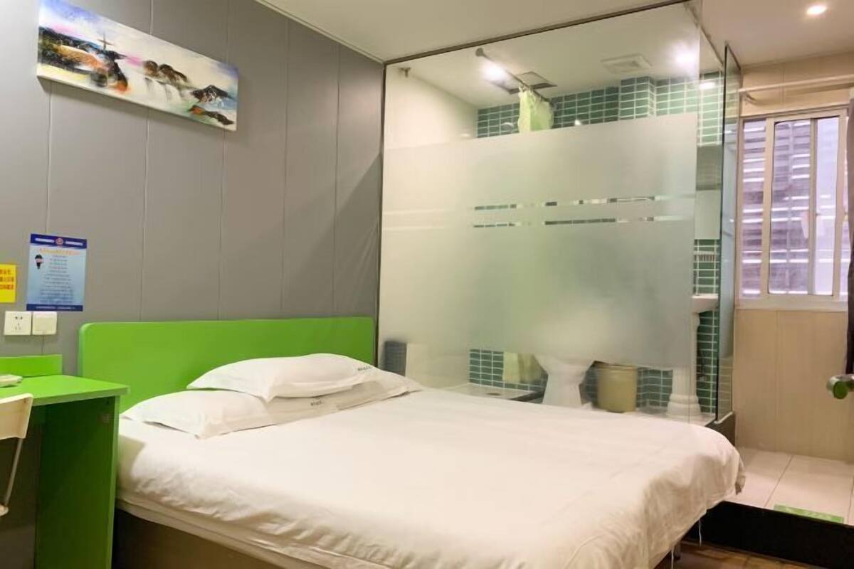 A hotel room with a green headboard, a bed with white bedding, a glass partition separating the room, and a window with blinds.