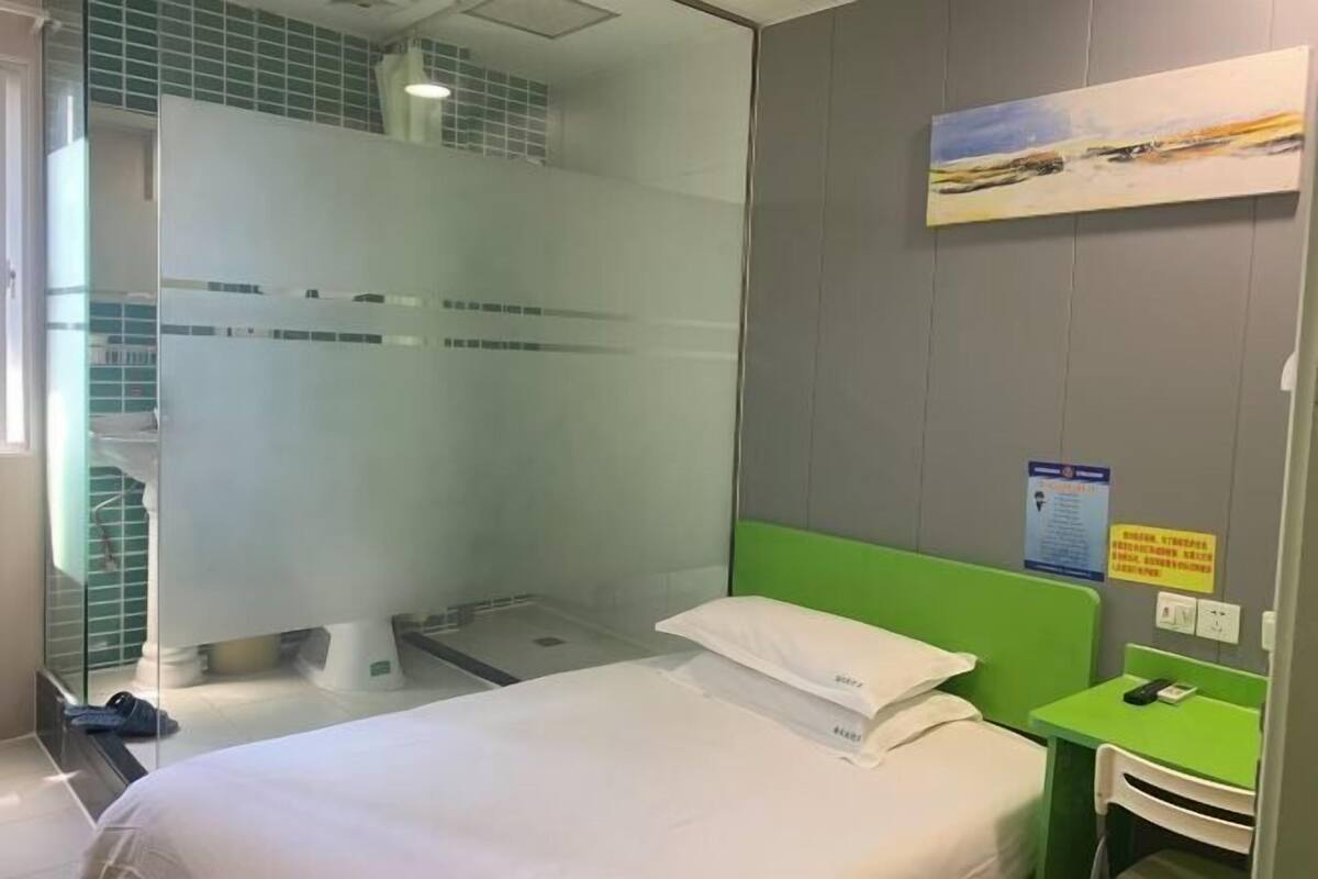 A hotel room with a bed, a green headboard, a glass shower enclosure, and a painting on the wall.