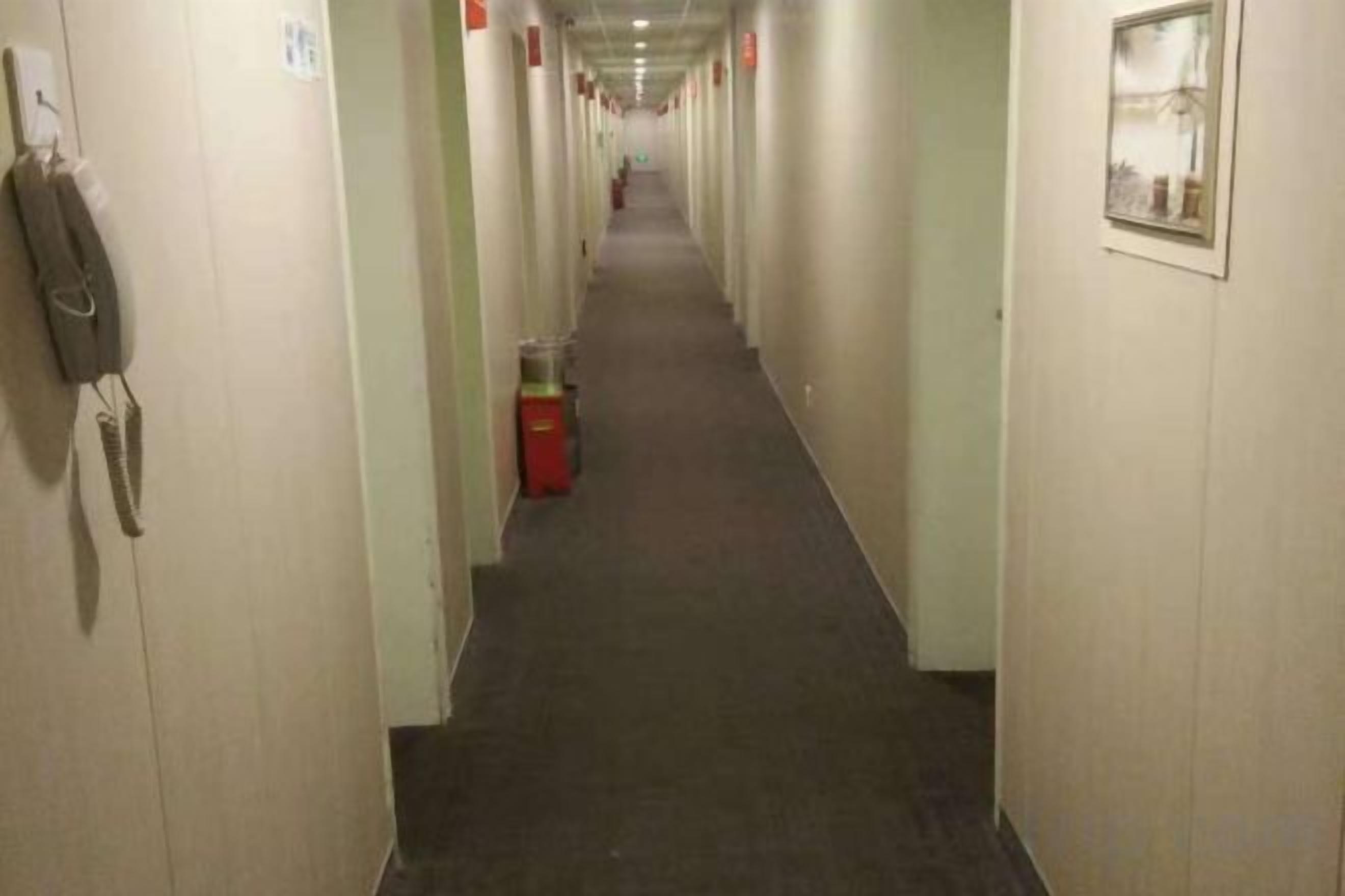 Un largo y estrecho pasillo de hotel con puertas a ambos lados, un bote de basura en el centro y un teléfono en la pared izquierda.