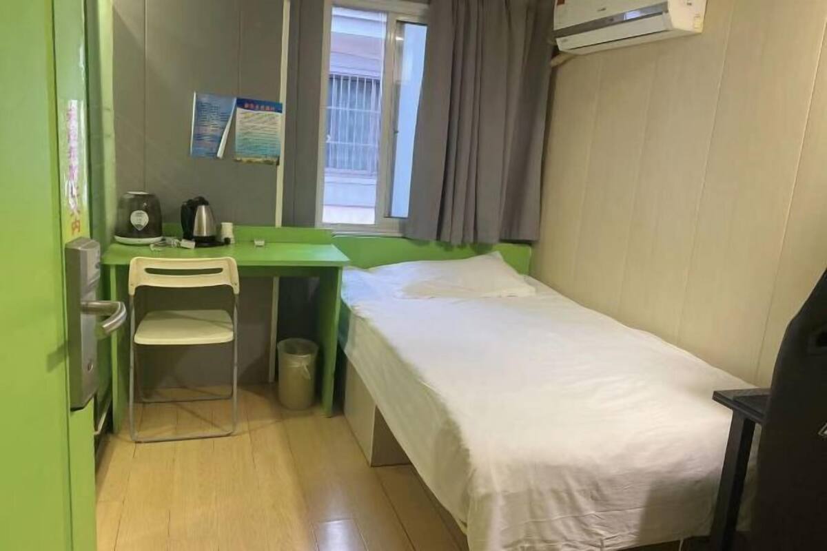 A small, single-bed room with a green desk, a chair, a kettle, and a window with curtains.