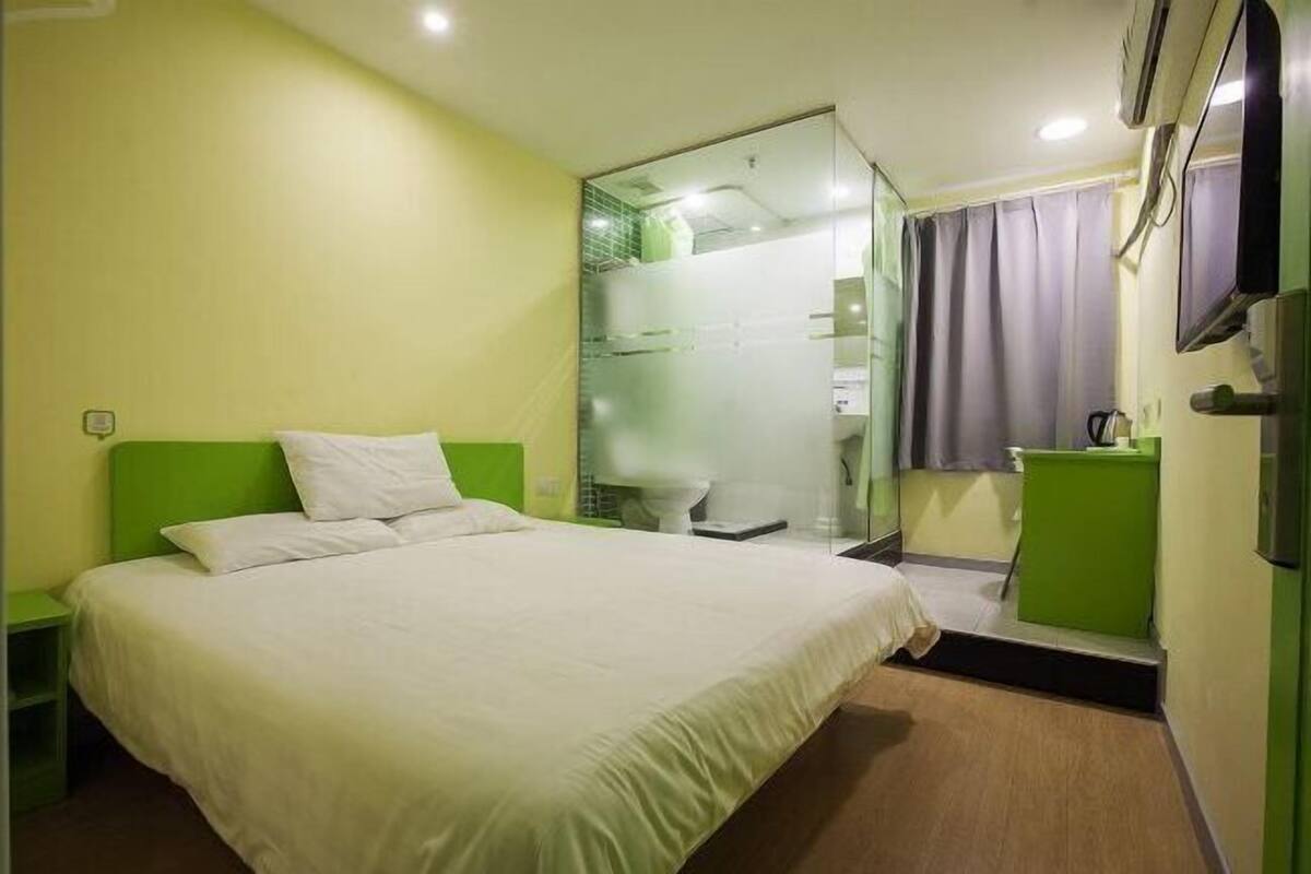 A hotel room with a bed, a green nightstand, a bathroom with a glass partition, and a mirror.