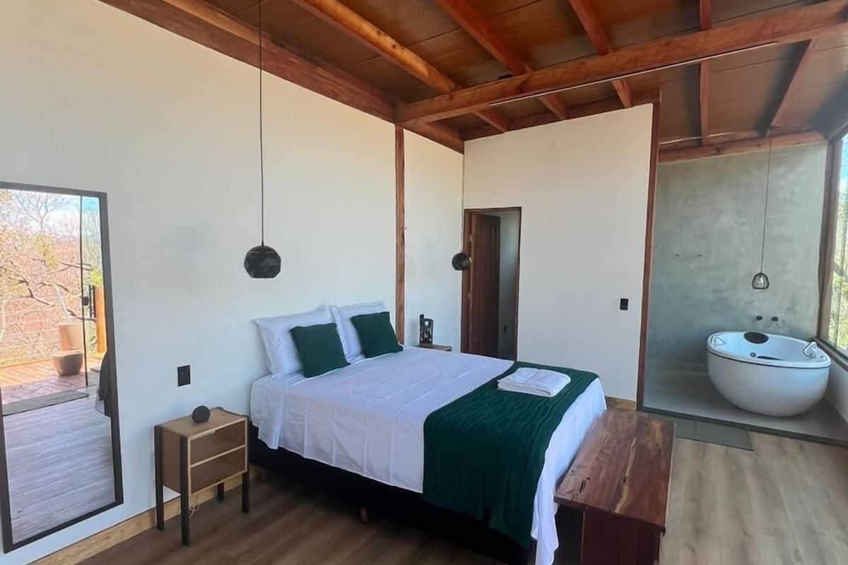 A bedroom with a wooden ceiling, a bed with white bedding and green pillows, a bedside table, a bench, a large window, and a bathroom with a bathtub.