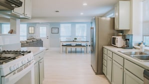 Een moderne keuken met witte kasten, een eethoek met een tafel en stoelen, en een uitzicht op een woonkamer met een bank en een raam.