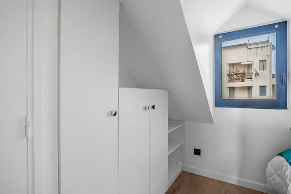 Una habitación con un armario blanco, un ventanal con vistas a un edificio y una cama con cubierta blanca.