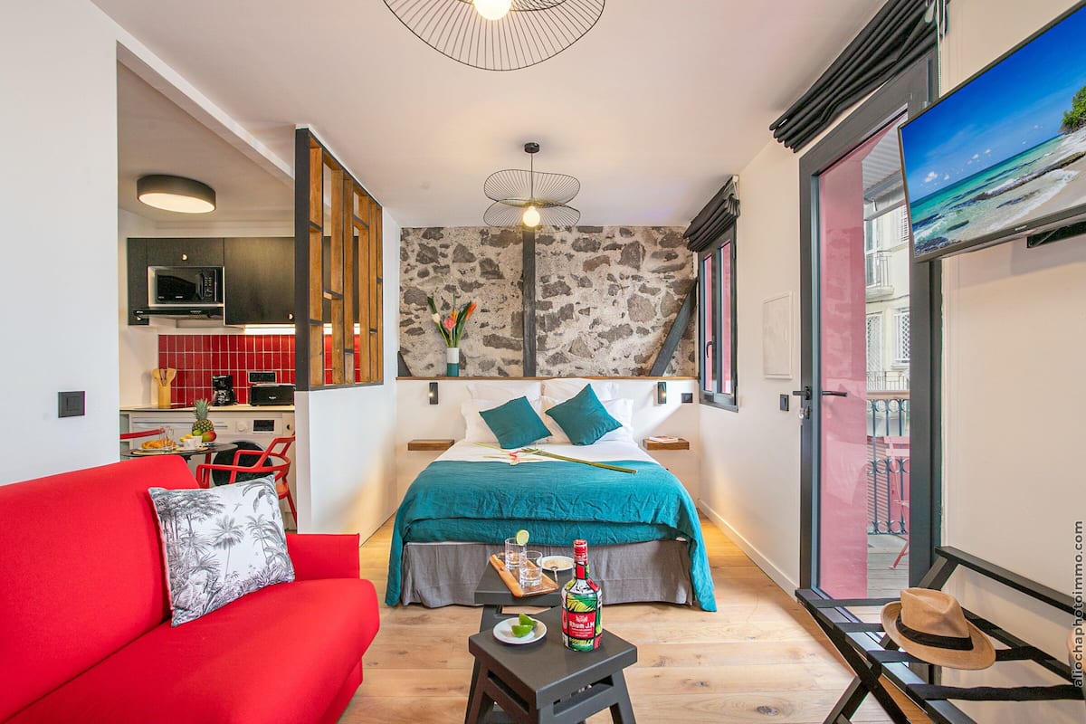 Un dormitorio compacto con cama, un sofá rojo, una mesita con una botella y copas, y un televisor en la pared.