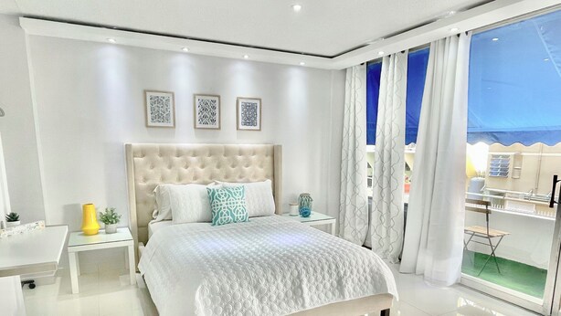 A modern bedroom with a large bed, a bedside table, a small desk, and a view of the outside.