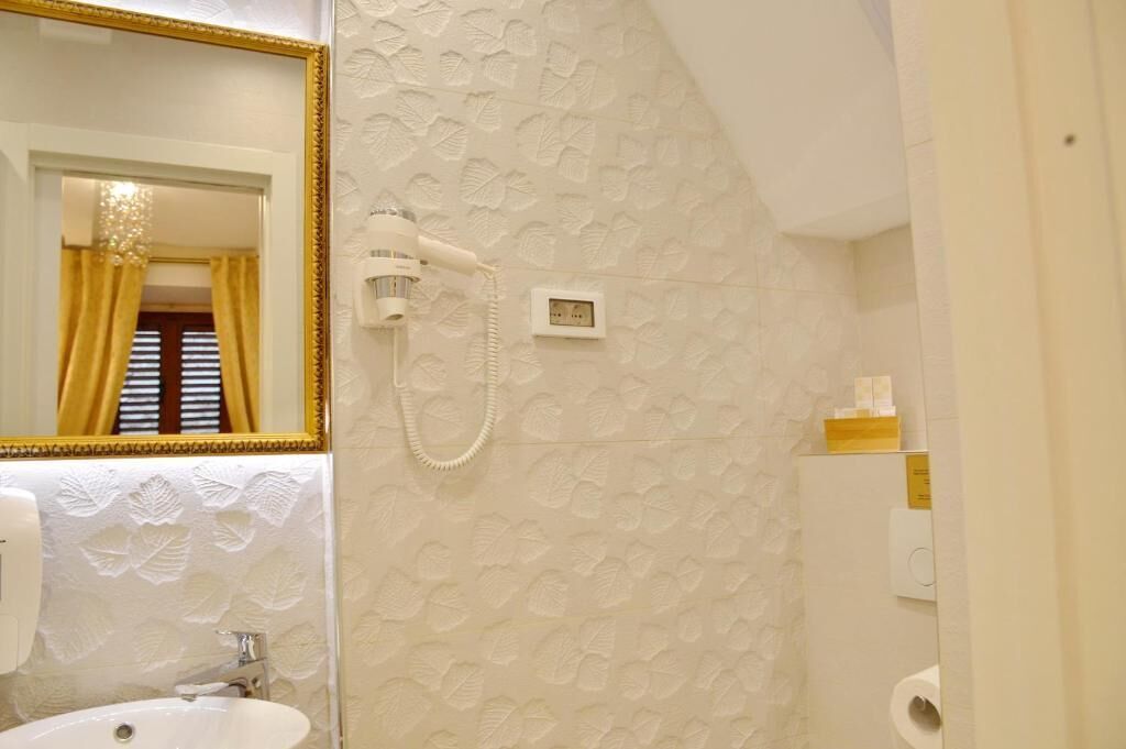 Une salle de bain avec un lavabo blanc, un miroir avec un cadre doré, un sèche-cheveux et un mur décoré d’un motif.