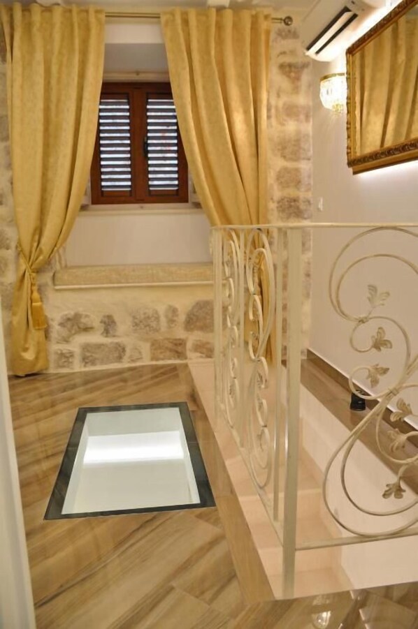 A room with a stone wall, a window with wooden shutters, a white metal bed frame, and a small bedside table with a lamp.