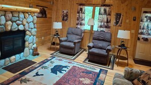 Een gezellig interieur van een blokhut met een stenen openhaard, twee fauteuils, een bank en een vloerkleed met dierenprint.