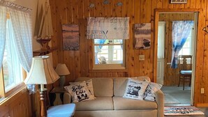 Een gezellig blokhutinterieur met houten details, een bank met decoratieve kussens, een tafel met een lamp en uitzicht op buiten.