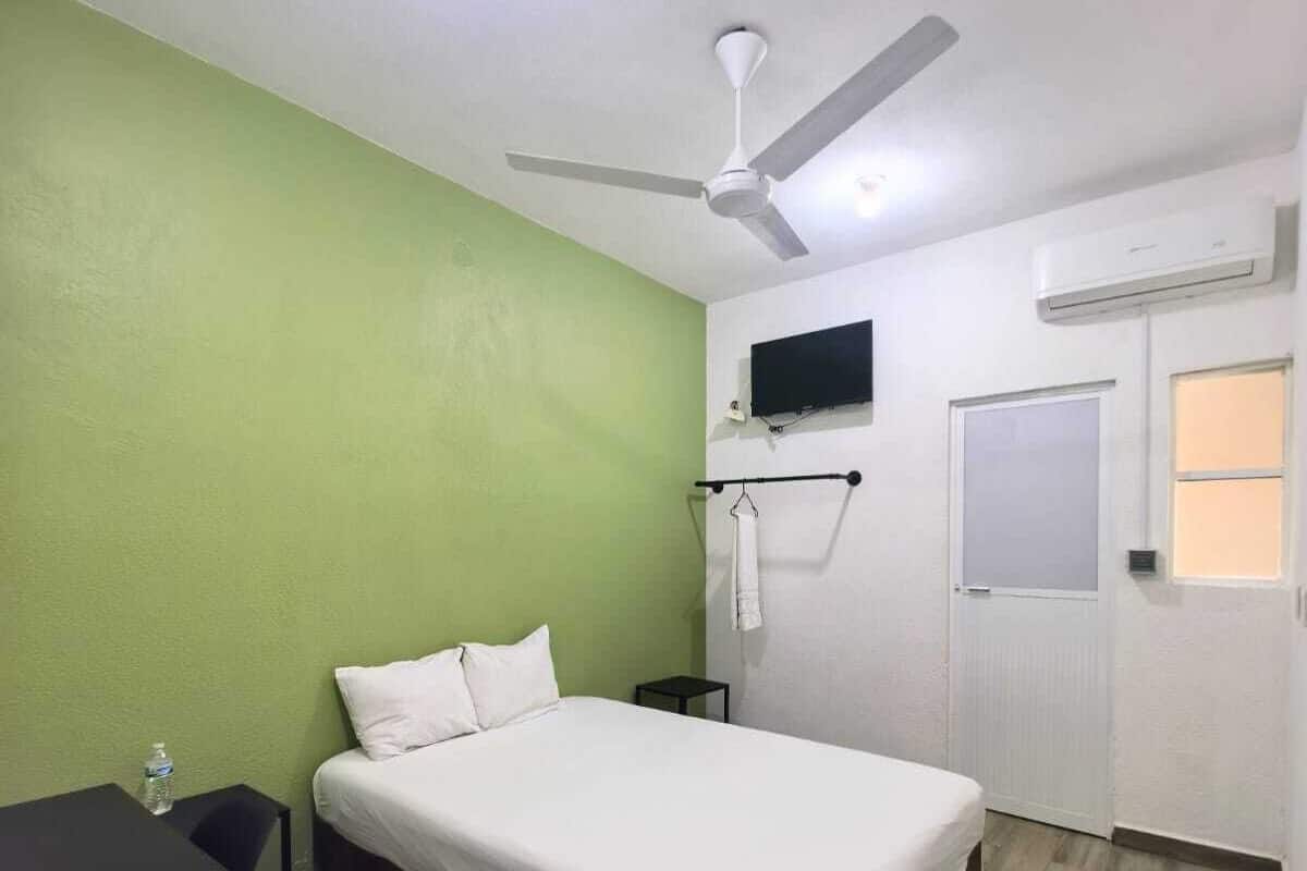 A bedroom with a bed, a ceiling fan, a TV mounted on the wall, and an air conditioning unit.