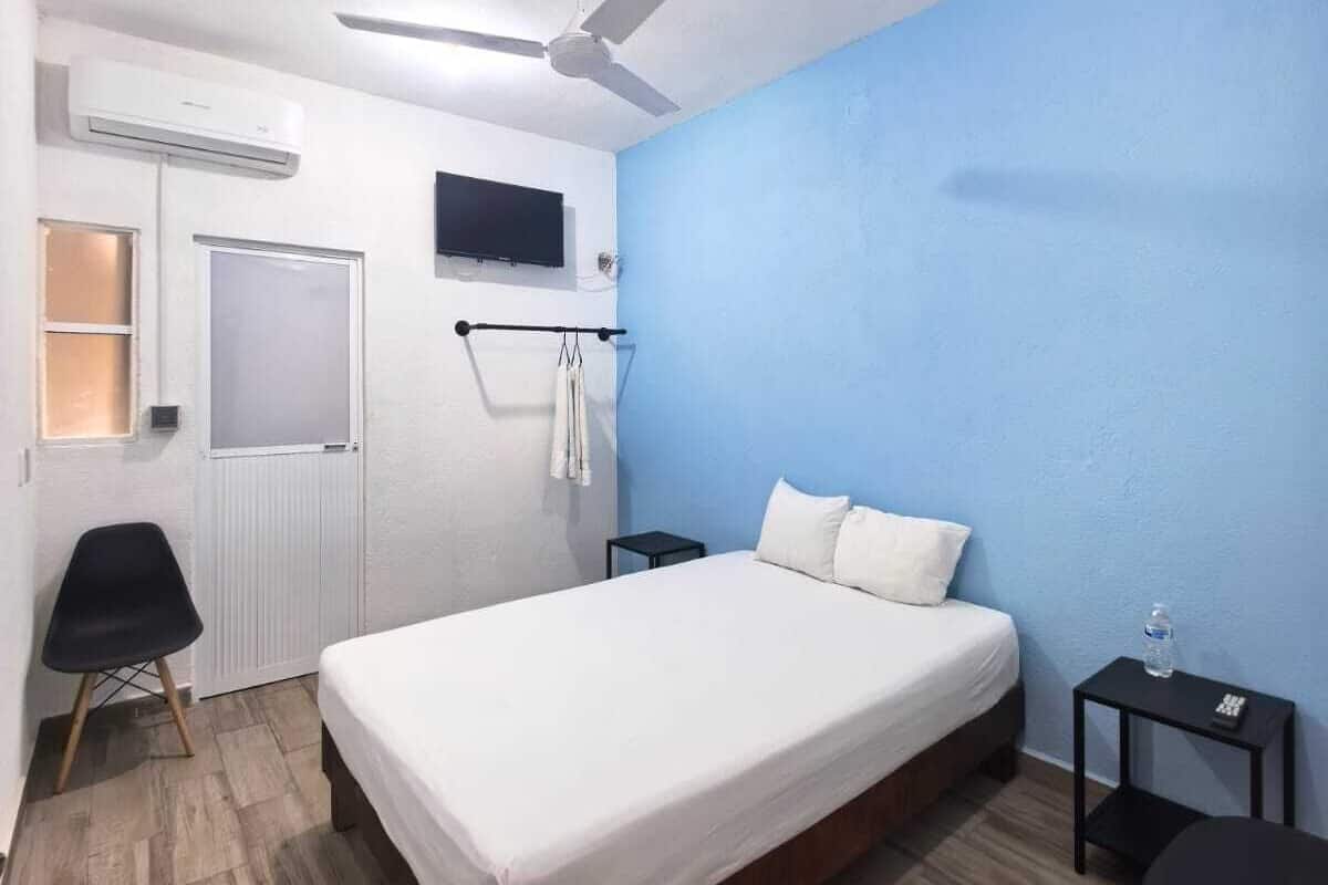 A bedroom with a bed, a ceiling fan, an air conditioning unit, a TV mounted on the wall, a small table, and a chair.