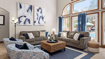 A living room with two sofas, a coffee table, and a patterned ottoman. There are abstract paintings on the wall and a view of a pool outside.