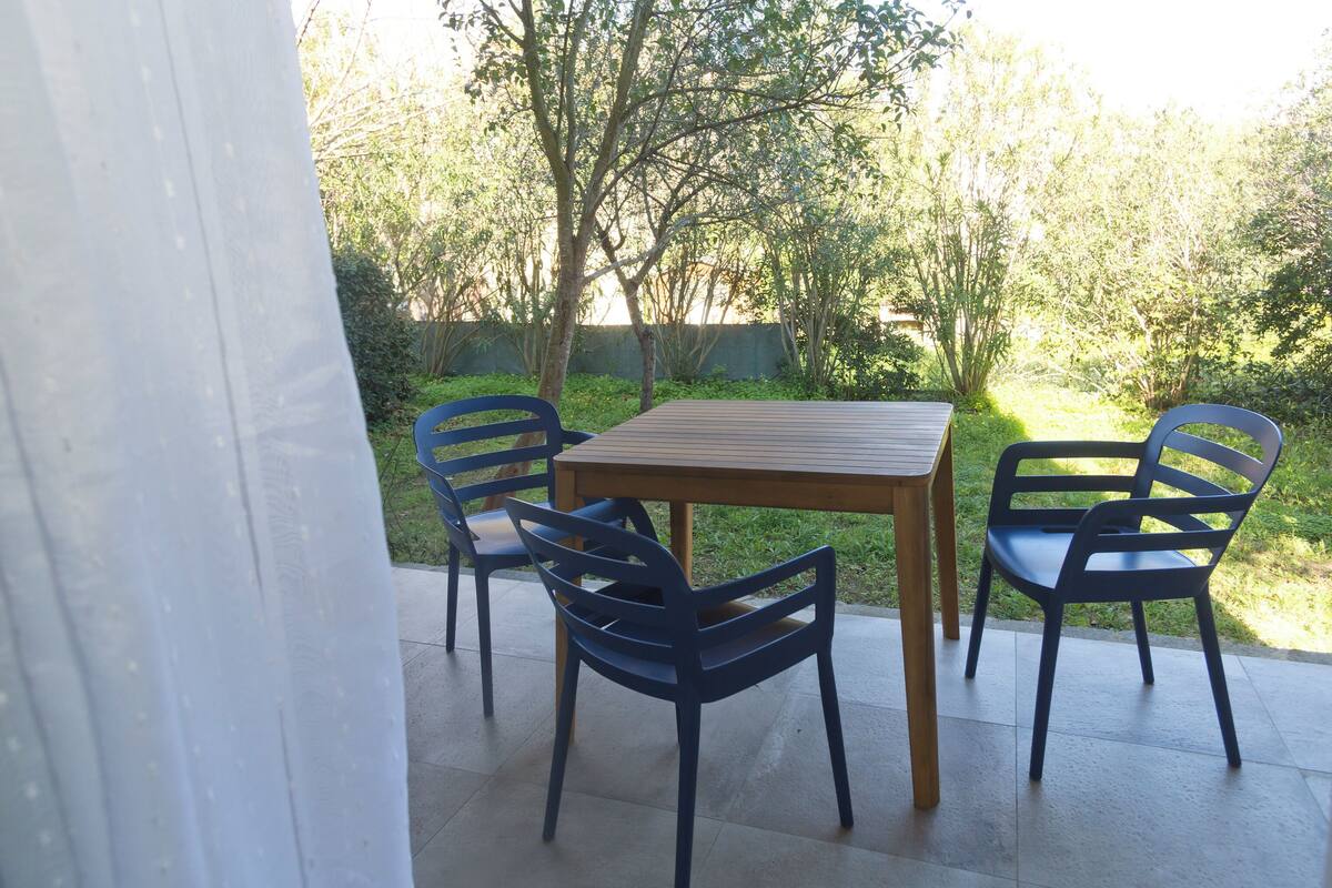 An outdoor patio with a wooden table and four chairs, surrounded by greenery and trees.