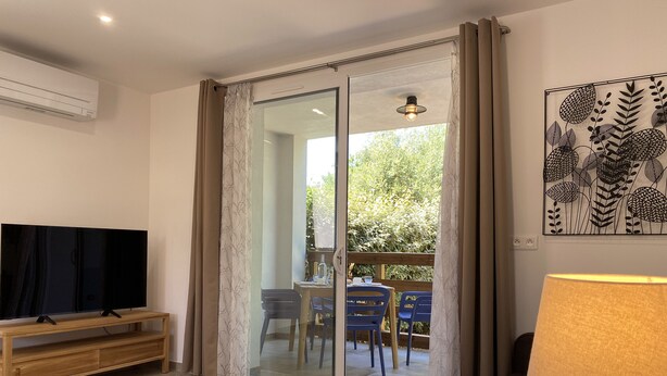A living room with a TV on a wooden stand, a sofa, a table, and a lamp. There is a sliding glass door leading to an outdoor area with a table and chairs. - Les Mûriers (Calvi)