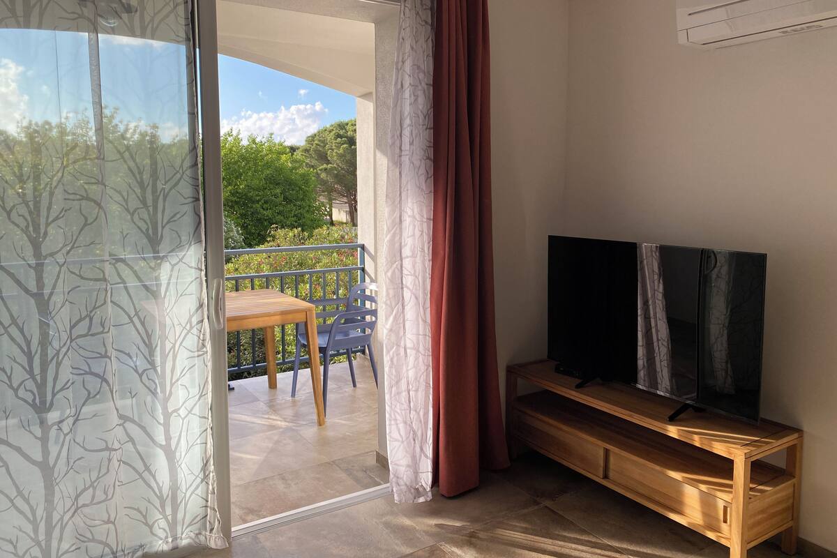 A room with a wooden TV stand, a balcony with a table and chairs, and a view of trees and greenery outside.
