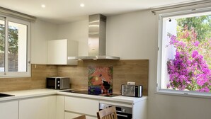 A modern kitchen with white cabinets, a stainless steel range hood, a microwave, and a toaster. There is a dining table with chairs and a window with a view of pink flowers. - Les Mûriers (Calvi)