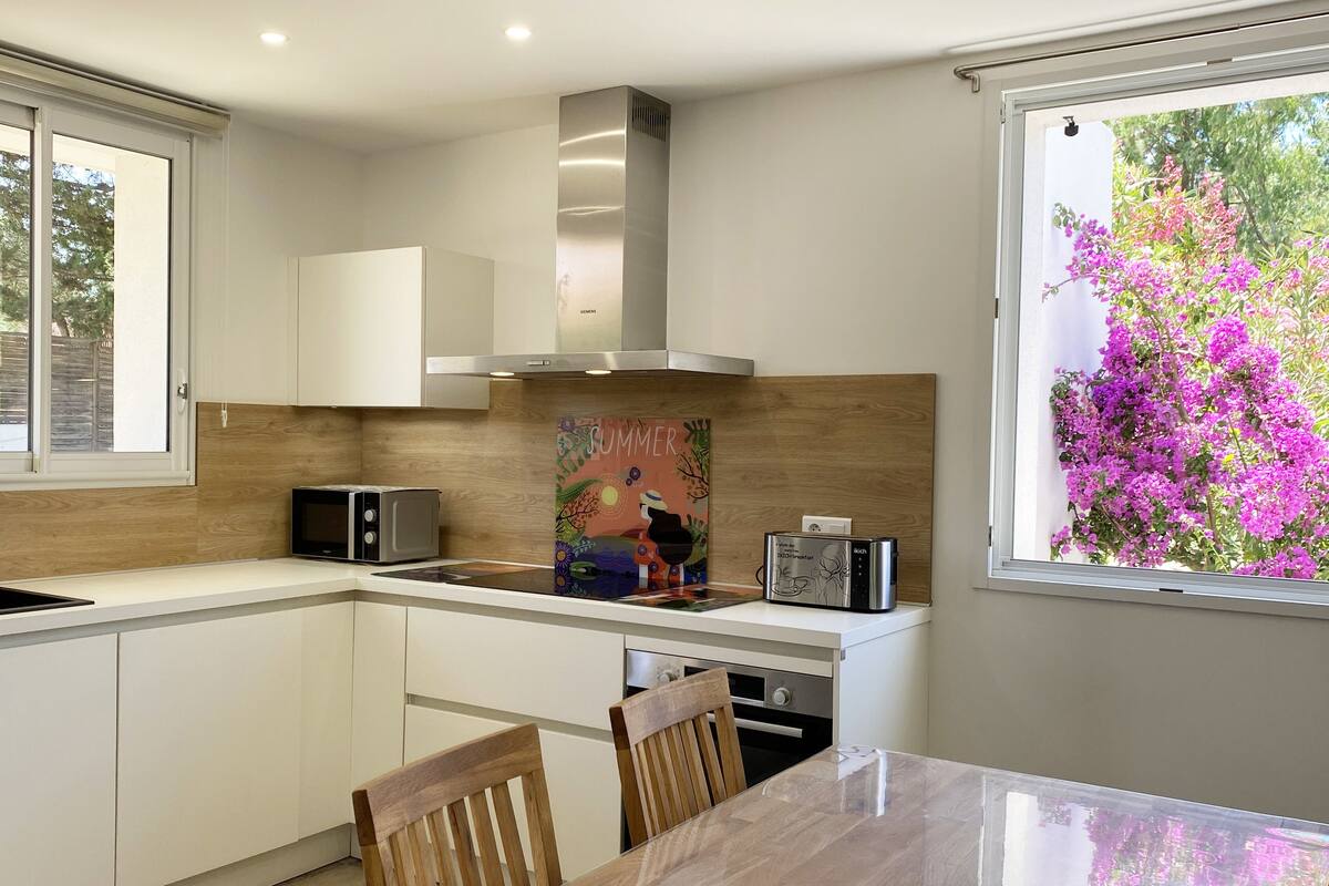 A modern kitchen with white cabinets, a stainless steel range hood, a microwave, and a toaster. There is a dining table with chairs and a window with a view of pink flowers.