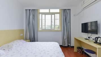 Quarto de hotel com cama, escrivaninha com televisão, janela com cortinas e uma cadeira.