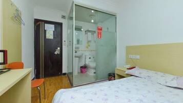 Quarto de hotel com cama, escrivaninha, cadeira, banheiro com vaso sanitário e pia, e porta de correr.