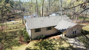 A two-story house with a sloped roof, surrounded by a wooded area and a small pond.