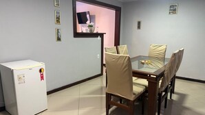 A dining area with a glass table and beige chairs, a white refrigerator, and a small TV on a shelf. - MARINAS DUPLEX HOUSE (Marinas Angra dos Reis)