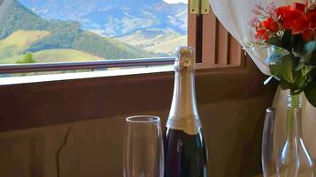 A table with a bottle of wine and two wine glasses, a vase with flowers, and a view of a mountainous landscape through a window with white curtains.