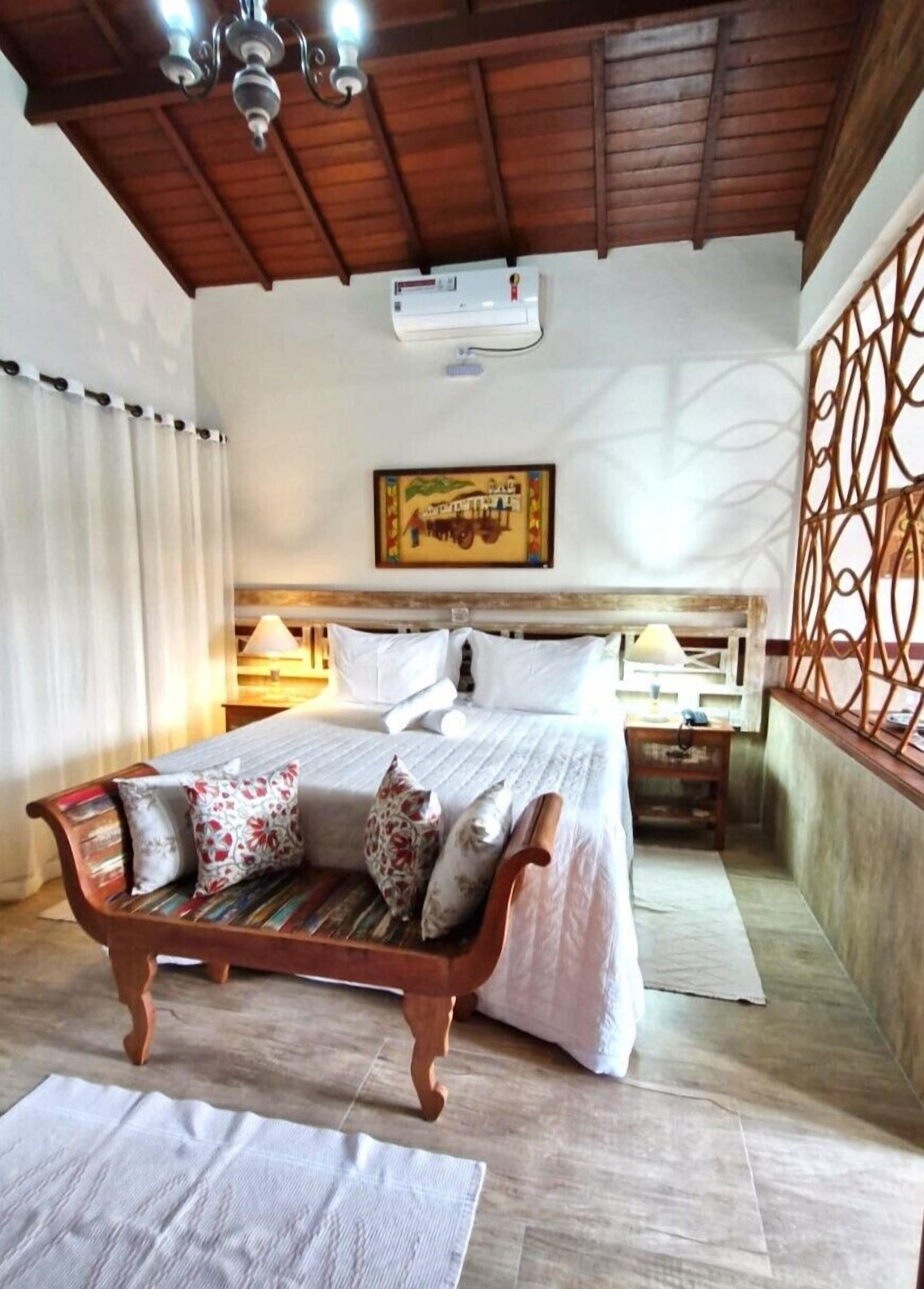 A bedroom with a wooden ceiling, a bed with white bedding, a bench with pillows, a nightstand with a lamp, and a decorative wall panel.