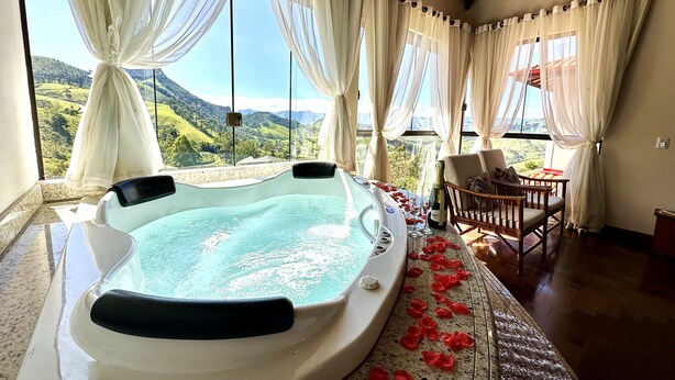A hot tub in a room with large windows, a view of green hills, and a wooden ceiling. - Aldeia Manacás Pousada Conceito (São Bento do Sapucaí)