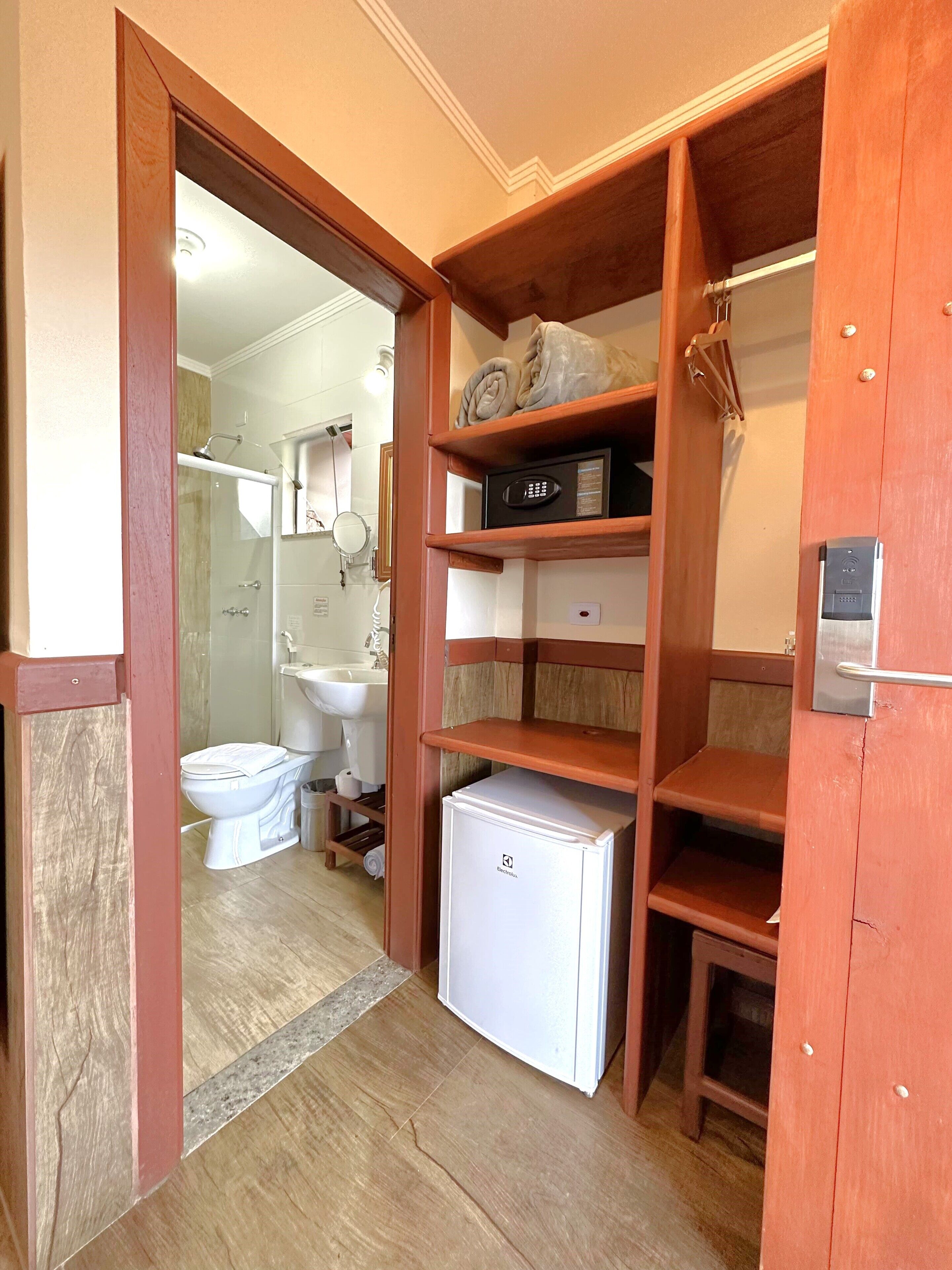 Baño compacto con inodoro, lavamanos y ducha. Incluye un refrigerador pequeño y una repisa con artículos de aseo.