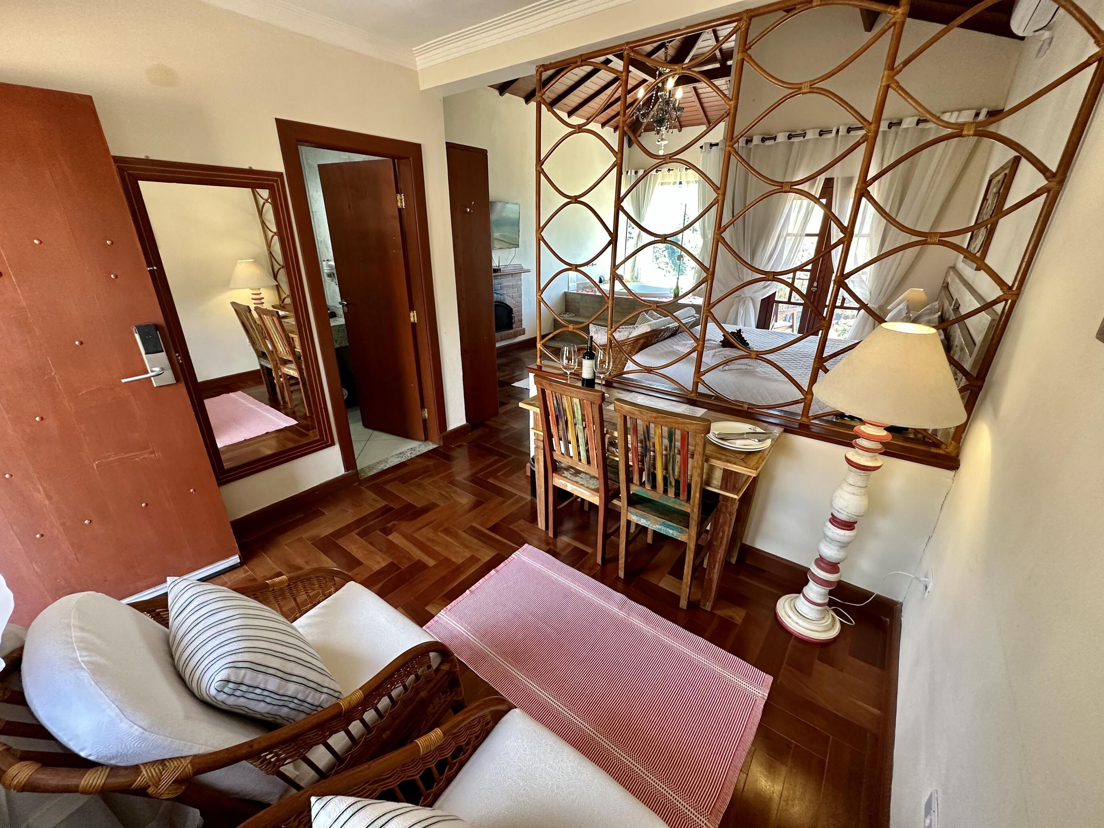 A cozy room with a wooden floor, a bed with a white bedspread, a wooden chair with cushions, a small table with a wine glass, and a decorative screen.