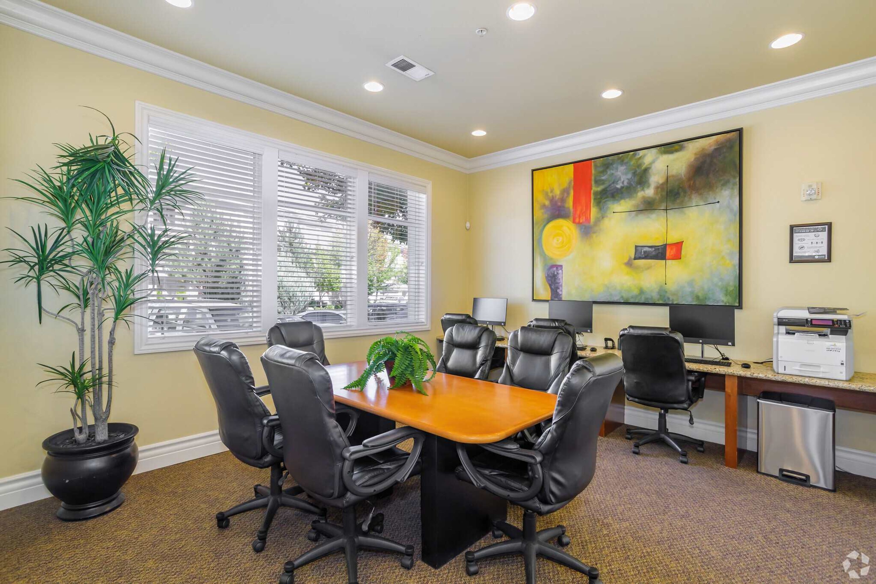 A conference room with a wooden table, black office chairs, a potted plant, a painting on the wall, and a printer on the desk.