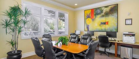 A conference room with a wooden table, black office chairs, a potted plant, a painting on the wall, and a printer on the desk.