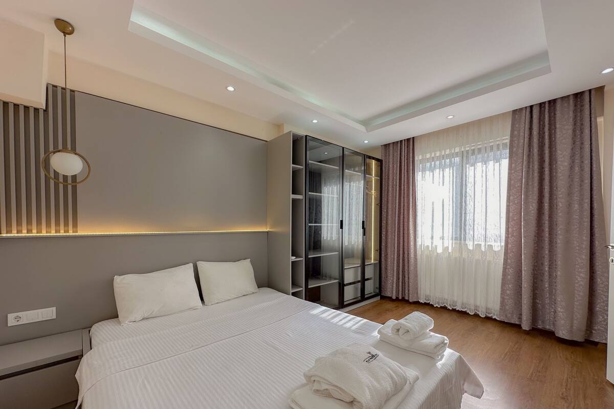 Ein modernes Hotelzimmer mit einem großen Bett, einem Kleiderschrank, einem Schminktisch und einem großen Fenster mit Vorhängen.