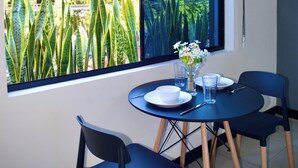 Et spiseområde med et bord dækket til to personer, et vindue der ud til grønne omgivelser, og en lille vase med blomster.