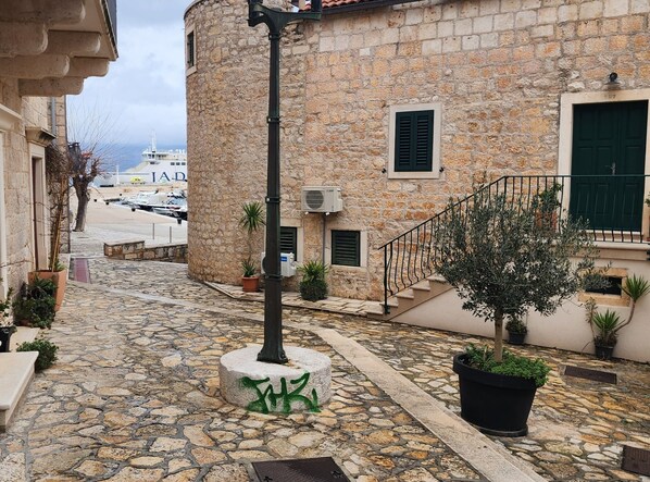 A stone building with a staircase leading to a door, surrounded by potted plants and a stone-paved street.