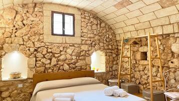 A stone-walled bedroom with a bed, two bedside tables, a window, and a wooden ladder.
