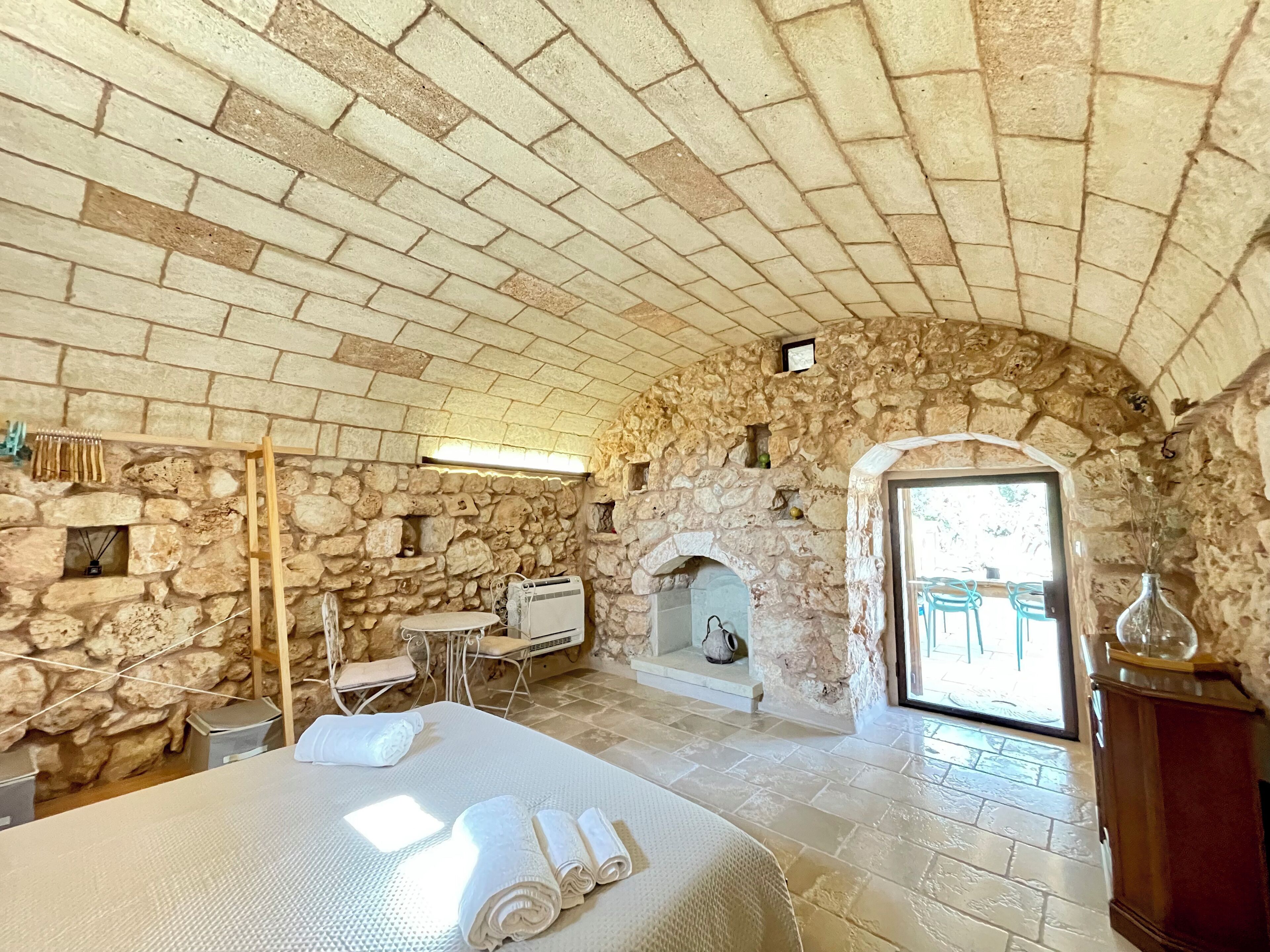 Une chambre aux murs de pierre, avec un lit, une petite table, une chaise, une cheminée et une vue sur l’extérieur.