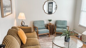 A living room with a patterned sofa, two armchairs, a round coffee table, a lamp, a mirror, and a window with blinds. - Charming 2-Bedroom Townhome in Downtown Cary (Cary)