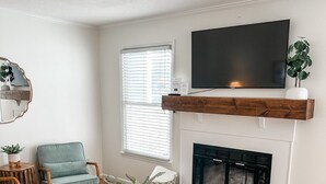 A living room with a fireplace, a television mounted above it, a round coffee table, and a blue armchair. - Charming 2-Bedroom Townhome in Downtown Cary (Cary)