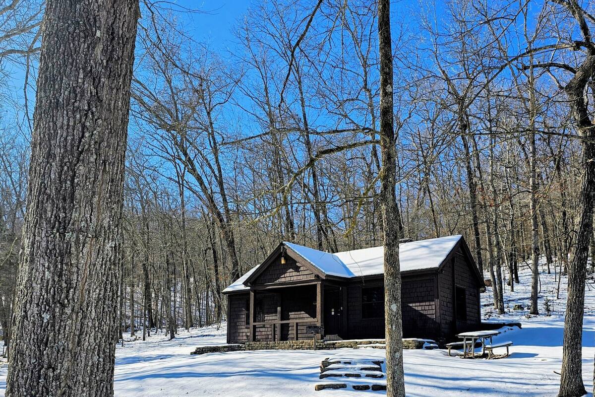 A rustic cabin with a stone foundation and wooden walls, surrounded by a snowy forest.