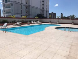 Piscina com espreguiçadeiras e uma pequena banheira de hidromassagem, cercada por um deck revestido.