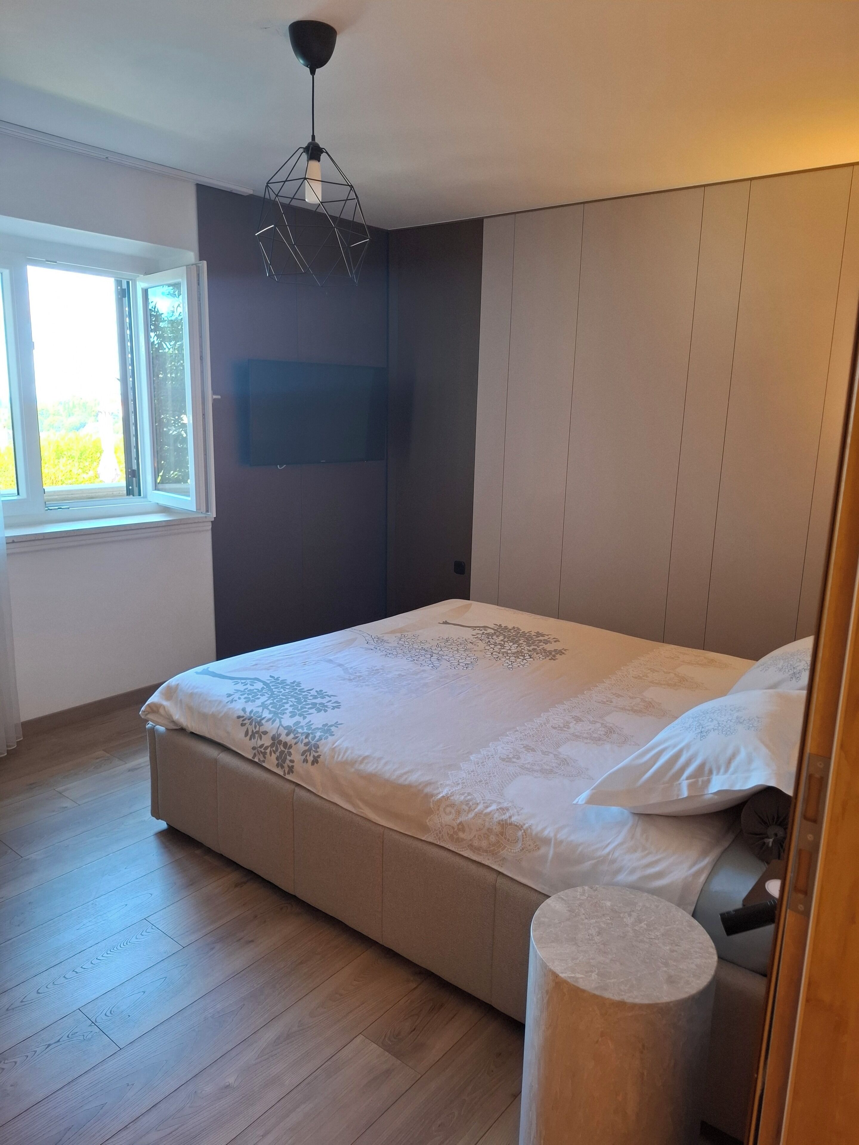 Een slaapkamer met een bed, een raam, een aan de muur bevestigde televisie en een houten vloer.