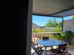 Een overdekt terras met een tafel en stoelen, en uitzicht op groen en gebouwen op de achtergrond.
